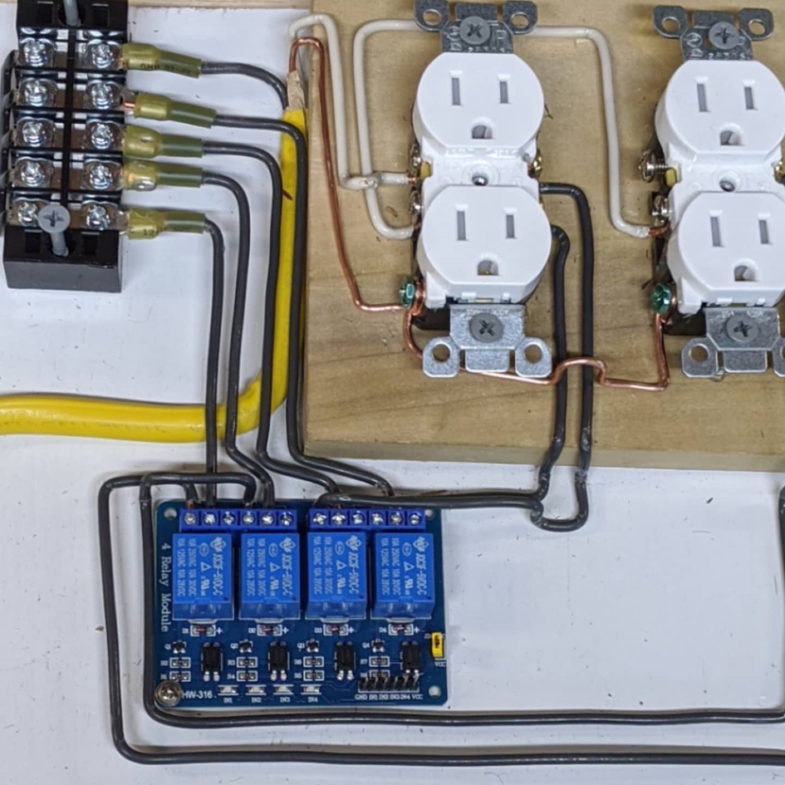 A mains switching board: a black barrier terminal block with crimped ring terminals on the left, a four-channel blue relay module below, and two white duplex receptacles mounted to plywood with copper ground bonding between them.