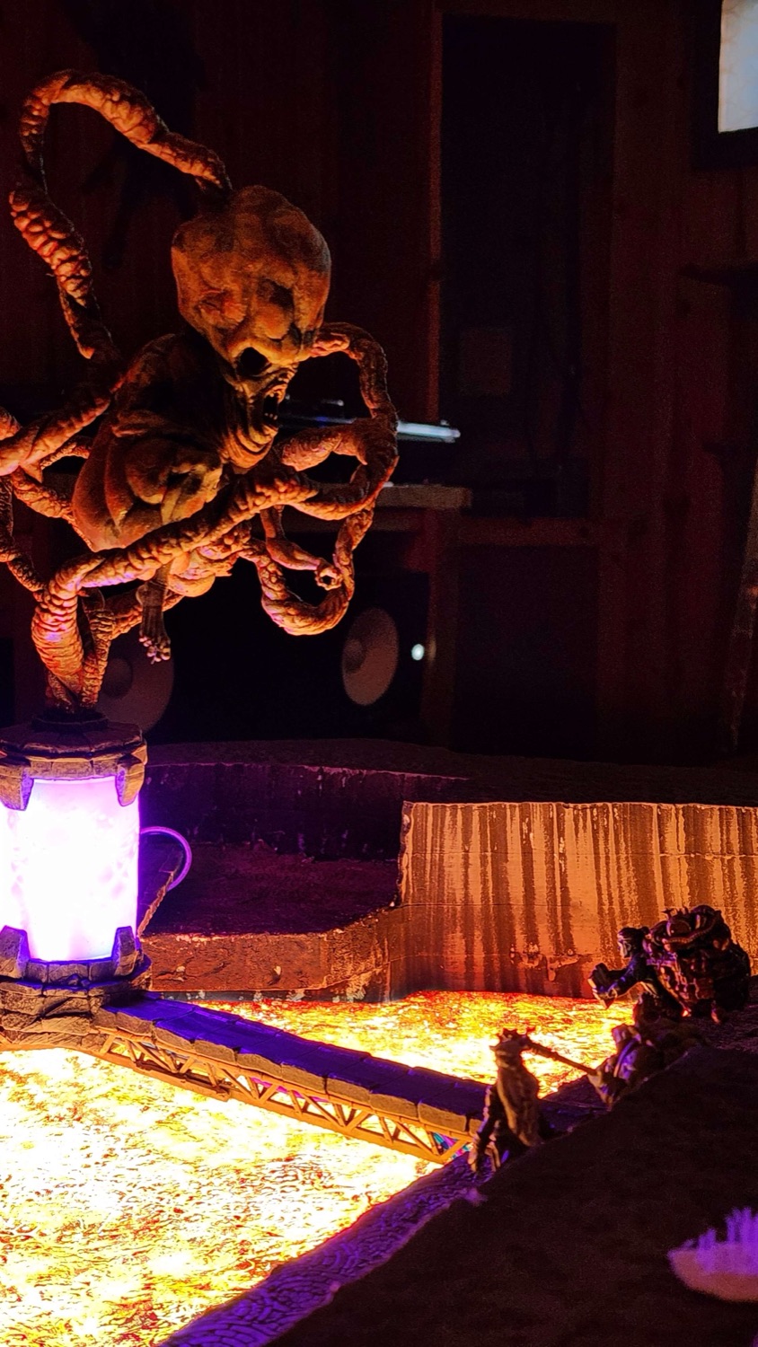 Low-angle night shot of a tentacled monster miniature mounted above a lit pillar, with the lava field glowing below and figures crossing a bridge.