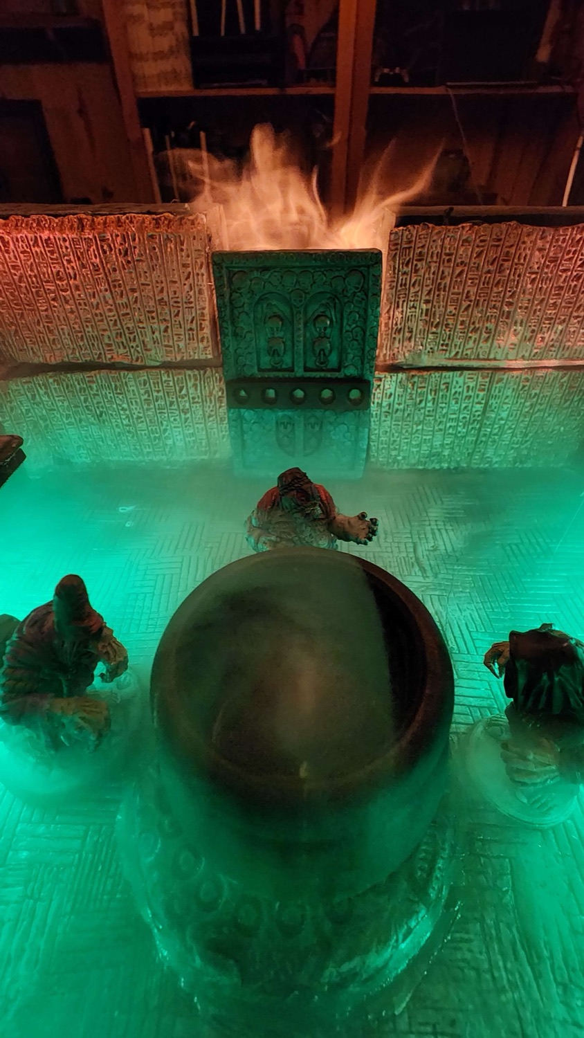 Close-up of the shrine platform lit in green, with fog rolling over a cauldron and painted miniatures around it.